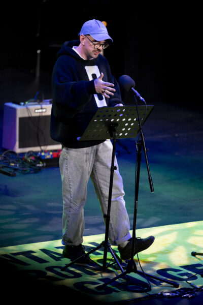 A man with a blue hat and glasses reads into a microphone on a stage - he is wearing a black jumper and jeans.