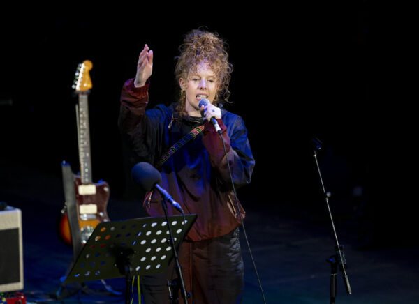 A woman with curly hair performs on stage - she is wearing a tracksuit that has a dark purple pattern and holds a microphone.