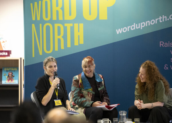 Four women sit on stage, one of them is talking -behind them is a big blue physical screen behind them that reads: Word Up North.