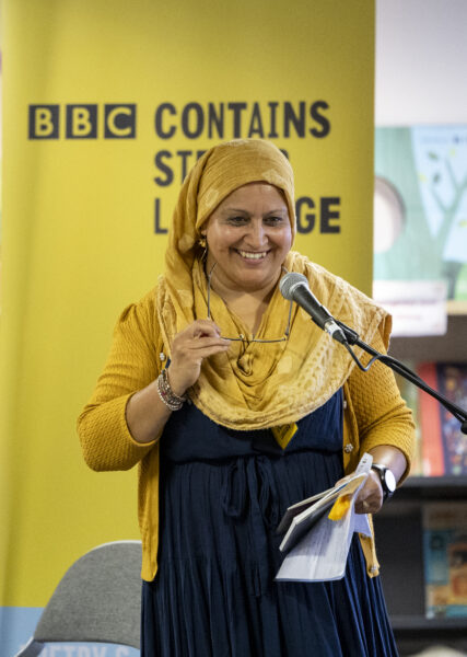 A woman wearing a yellow hijab smiles by a microphone - a yellow banner behinds reads: BBC Contains Strong Language.