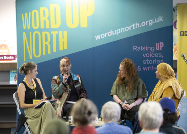 Four women sit on stage, one of them is talking -behind them is a big blue physical screen behind them that reads: Word Up North.