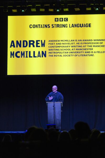 A man stood on a stage talking, with his name on a large bright yellow screen behind him 'Andrew McMillan'