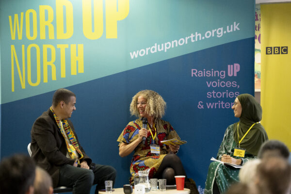 Three people sit on the stage before a crowd - a big blue physical screen behind them that reads: Word Up North.