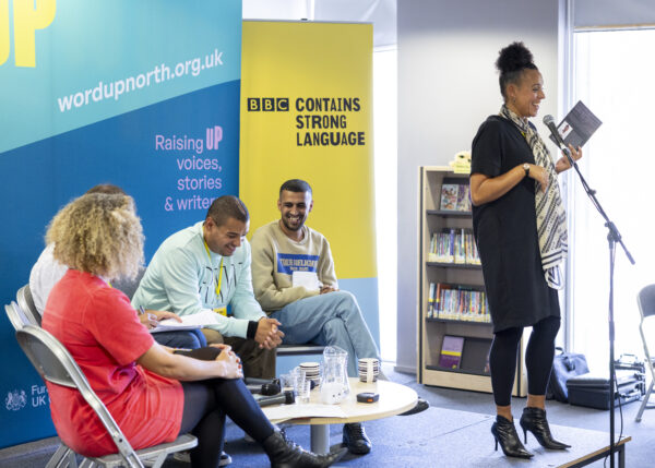 A woman with dark curly hair reads into a microphone, four people sat behind her on stage.