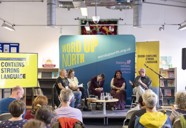 Five people sit on stage before a crowd big blue physical screen behind them that reads: Word Up North.