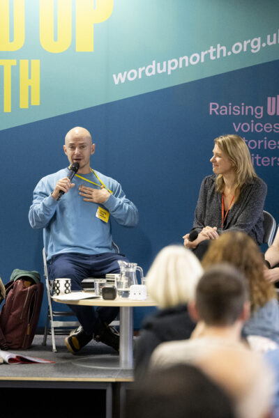 A man wearing a blue sweatshirt speaks into a microphone.