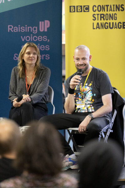 A man wearing a black t-shirt speaks into a microphone.