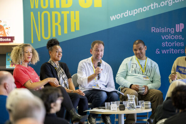A man talks into a microphone - he is on stage with four other people and they are sat before a big blue physical screen behind them that reads: Word Up North.