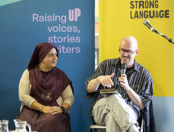 A bald man wearing glasses speaks into the microphone, a woman in a burgundy hijab looks at him as he speaks.