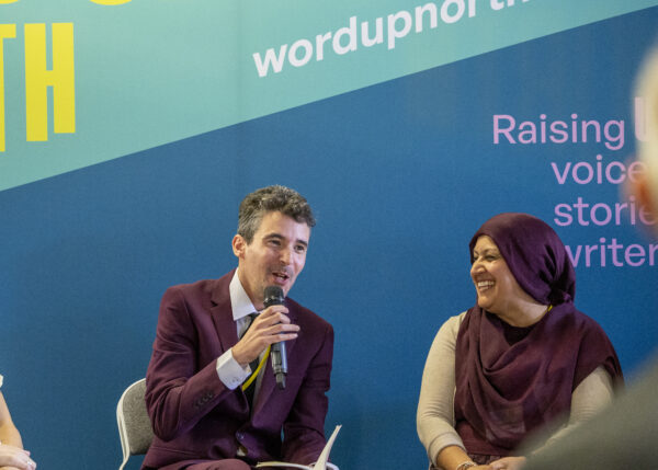A man in a burgundy suit speaks into the microphone - a woman in a burgundy hijab smiles at him as he speaks.