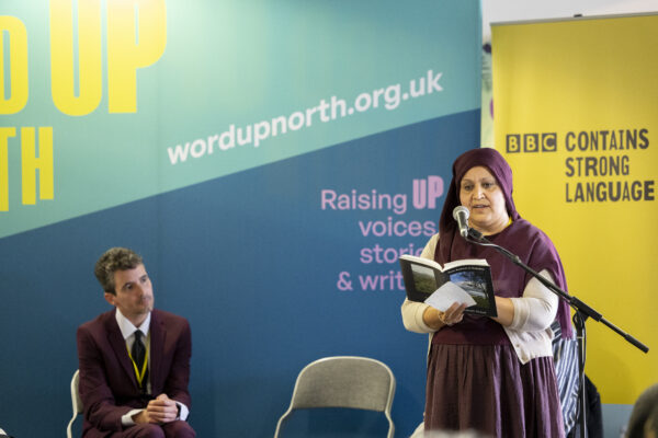 A woman in a burgundy hijab reads from a book into a microphone.