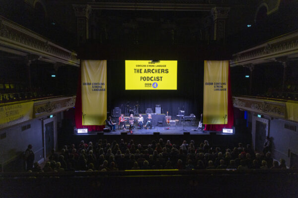 The stage of St. George's hall is set for a live broadcast with lots of equipment, and is dressed with yellow banners - the screen reads: The Archers Podcast.