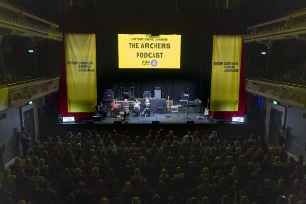 The stage of St. George's hall is set for a live broadcast with lots of equipment, and is dressed with yellow banners - the screen reads: The Archers Podcast.