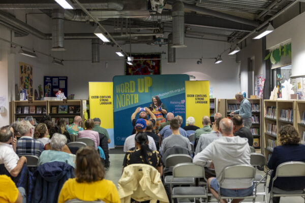A woman reads in front of an audience - behind her is signs reading: Word Up North and BBC Contains Strong Language.