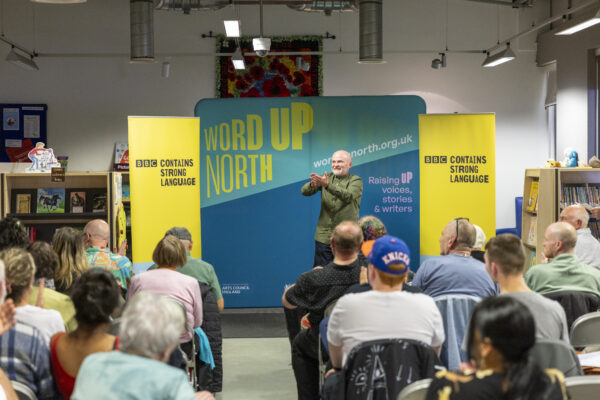 A man in a green shirt reads in front of an audience - behind him is signs reading: Word Up North and BBC Contains Strong Language.