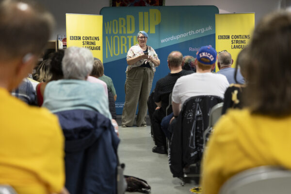 A person reads in front of an audience - behind them is signs reading: Word Up North and BBC Contains Strong Language.