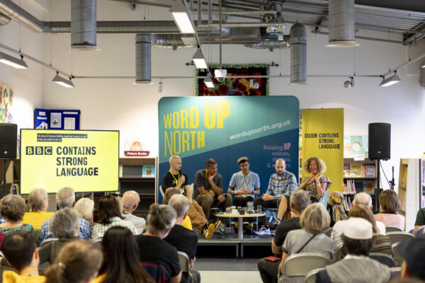 Five people sit on the stage before a crowd - a big blue physical screen behind them that reads: Word Up North.