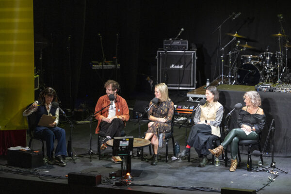 Four people are sat n stage, three women and one man, each with big microphones in front of them - they are looking to the woman on the far left who is reading.