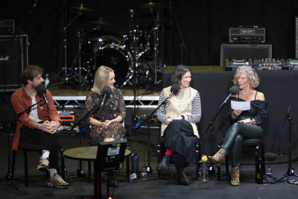 Four people are sat n stage, three women and one man, each with big microphones in front of them - they are looking to the woman on the far right who is pulling a face.