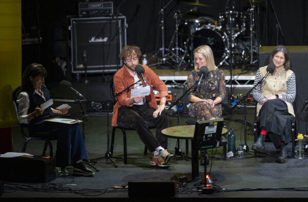 Four people are sat n stage, three women and one man, each with big microphones in front of them - they are looking to the men, who is reading.