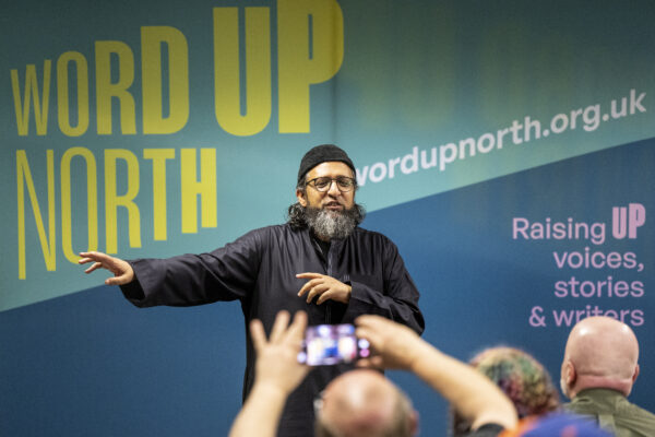 A man gestures as he performs to an audience - A man in a flatcap and green shirt reads in front of an audience - behind him is signs reading: Word Up North and BBC Contains Strong Language.