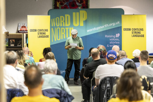 A man in a flatcap and green shirt reads in front of an audience - behind him is signs reading: Word Up North and BBC Contains Strong Language.