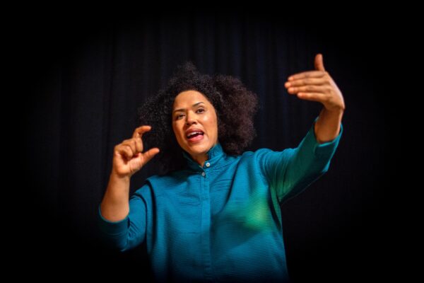 A black woman with curly hair poses for the camera - she is wearing a blue top and the background is black fabric.
