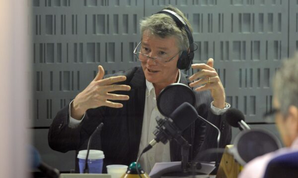 A white man talks into a microphone, he is wearing a white shirt and a black blazer and has a pair of headphones on his head.