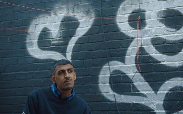 A south asian man poses for the camera in front of a grey wall decorated with white spray-painted hearts.