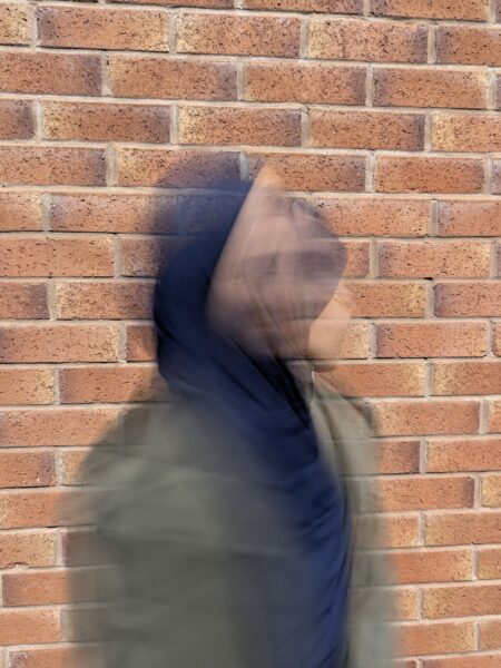 A layer photo of blurry figures, wearing a green jacket and a blue headscarf, overlaid against a brick wall.