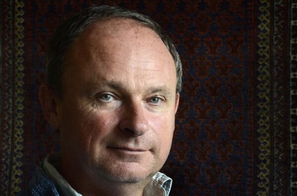 A man looks into the camera - he is wearing a collared shirt and the background is dark