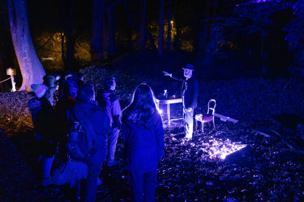 a ghostly looking man dimly lit, talking to a group of people in a dark woods