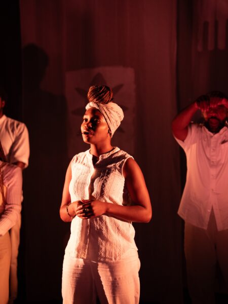 A black woman with her hair tied up in a scarf stands on stage.