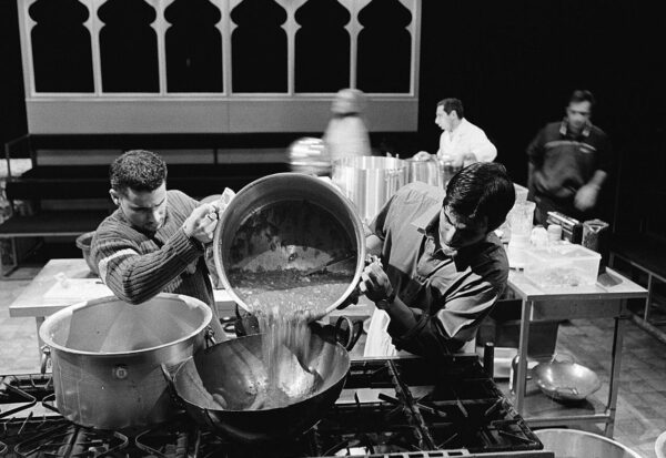 Tow men in a kitchen pooring a sauce out of a large pan into a hot wok