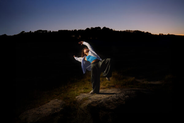 A dancer poses in the dark just before dawn. They are outside, wearing a blue top, jacket and loose trousers.