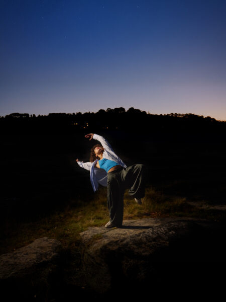 A dancer poses in the dark just before dawn. They are outside, wearing a blue top, jacket and loose trousers.