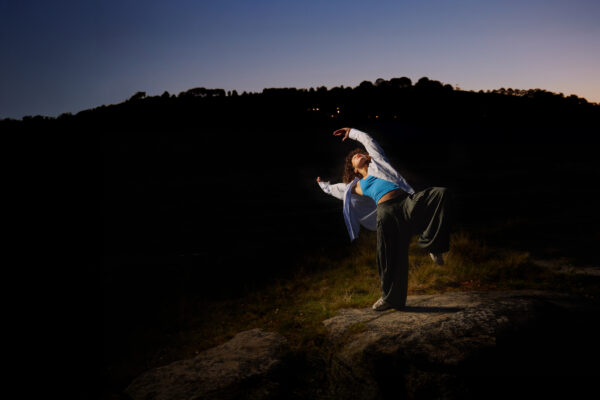 A dancer poses in the dark just before dawn. They are outside, wearing a blue top, jacket and loose trousers.