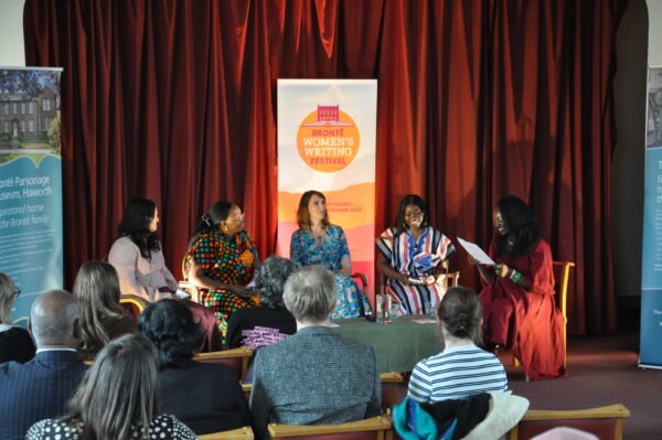 Five women sit at the front of an audience - there are four black women and one white woman in the middle. There is red curtains behind them and a banner reading: Bronte Women's Writing Festival. The woman on the right is reading.