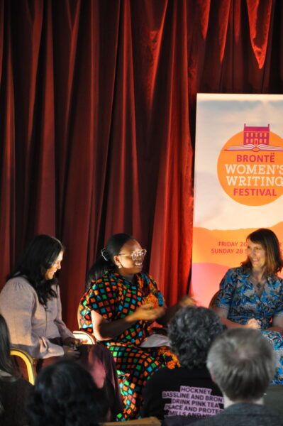 A close up shot - three women sit at the front of an audience - there are two black women and one white woman on the right. There is red curtains behind them and a banner reading: Bronte Women's Writing Festival. The woman in the middle is speaking.