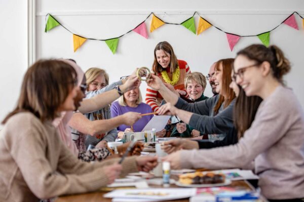 A group of people sit around a table smiling. They are passing pencils across the table.