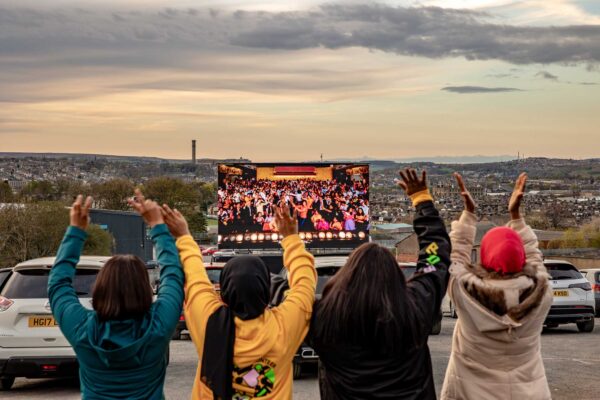 Four people raise their hands in the air in front of an outdoor cinema screen, on a hill high above Bradford.