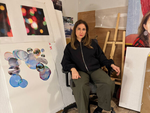 Artist Faiza Butt sits in a studio. She has artworks on paper around her and an easel.