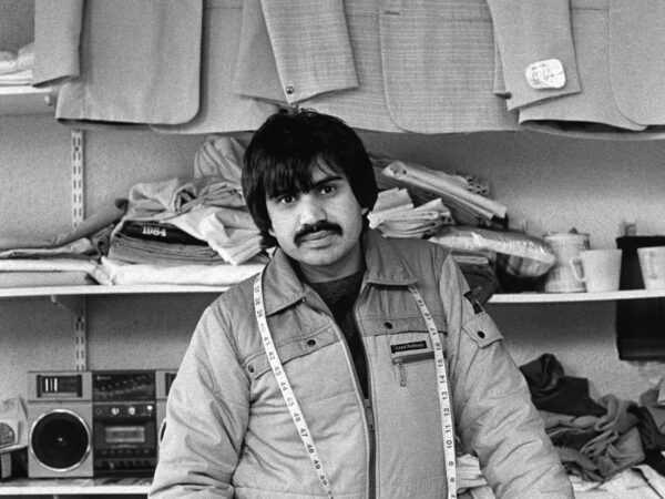 A black and white photograph of a man standing in a tailor's shop, looking towards the camera. He has a tape measure around his neck.