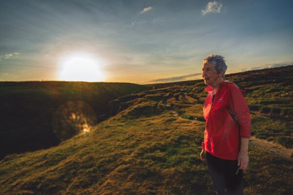 A woman with short grey hair in a red lightweight jacket stands on a moor with the sun rising in the distance.