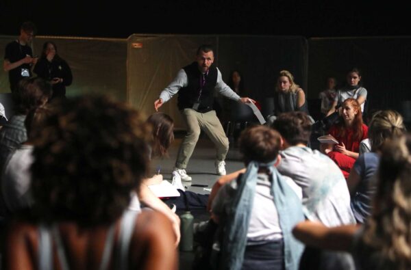 A man stands in a semi crouched position in front of a group. He appears to be leading a workshop or rehearsal.