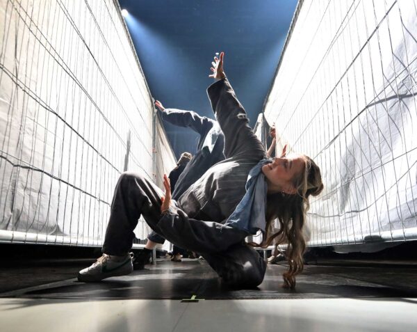 A dancer kneels close to us, in a narrow alley created by two tall white fences.