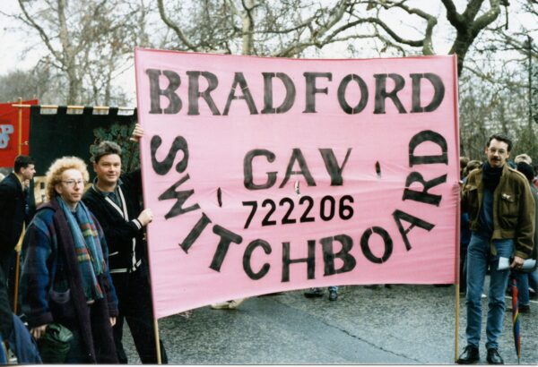 A digitised photo depicting three white men posing with a big pink banner - the black text reads: BRADFORD GAY SWITCHBOARD 722206.