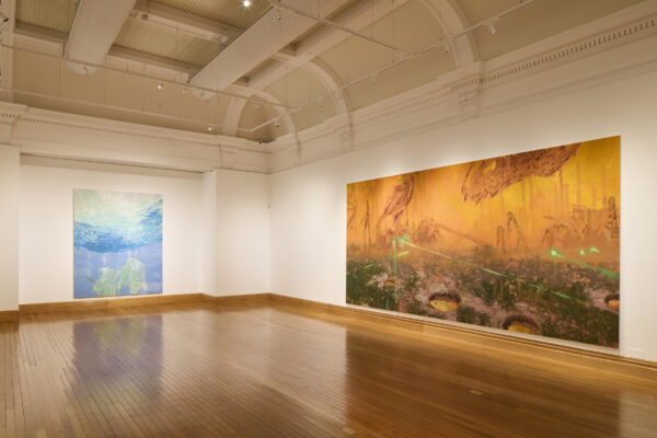 An installation view of Mohammed Sami's work in Cartwright Hall Art Gallery. In a large room with a wooden floor we see two paintings, one in shades of blue and another in oranges, yellows, and grey.