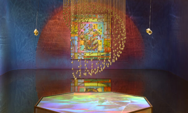 An artwork hanging from the ceiling in a gallery. The work has lots of small gold objects hanging on long strings. Behind this there is a painting on the wall with bright colours.