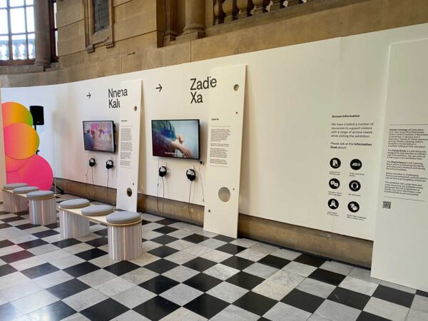 A white wall with two TV screens on it. Next to the screens are headphones and some small seats.